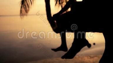 在令人惊叹的日落中，年轻幸福夫妇坐在棕榈树上的剪影。 挥舞着双腿。 双脚合拢